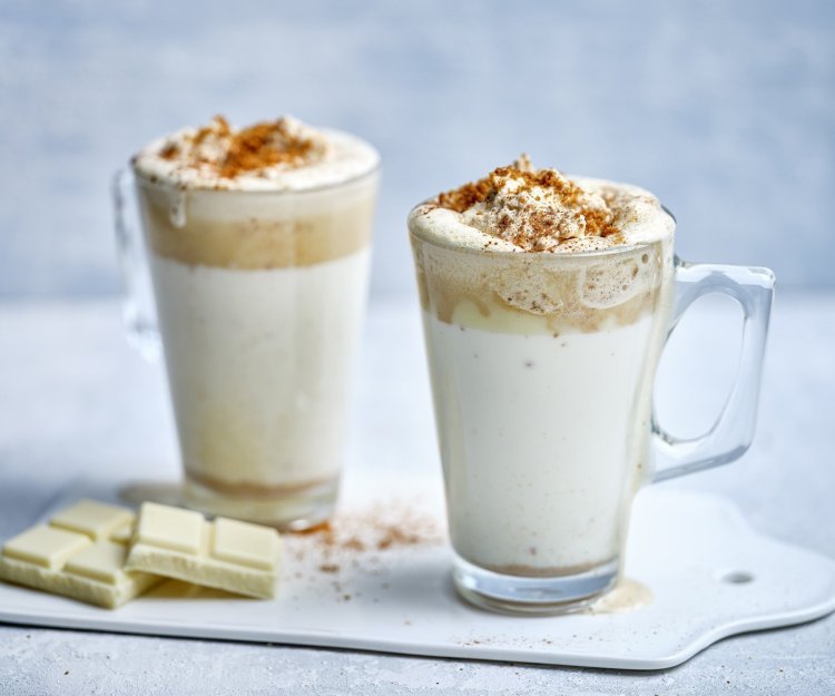 Twee hoge glazen tassen staan op het aanrecht klaar om te serveren. Ze zijn gevuld met witte chocolademelk, een goeie toef speculaasroom er bovenop en wat speculaaskruim. Er ligt een stukje witte chocolade naast. 