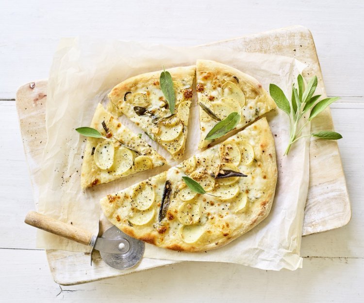 Witte pizza met aardappelplakjes, mozzarella en salie Een pizza belegt met mozzarella, plakjes aardappel en salie ligt op een wit boterpapiertje. Er ligt een pizzasnijder langs de pizza en nog enkele verse blaadjes salie.