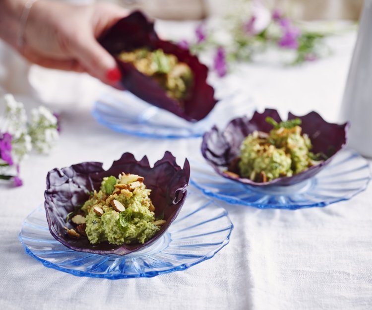 Wrap van rode kool met erwtenhummus en gerookte amandelen Er liggen 3 wraps van rode koolbladeren, elk op een blauw kristallen bordje. De wrap is gevuld met hummus van groene erten en geroosterde en gerookte amandelen. Een vrouwenhand neemt alvast een wrap tot haar hapje, geen tijd te verspillen dus.