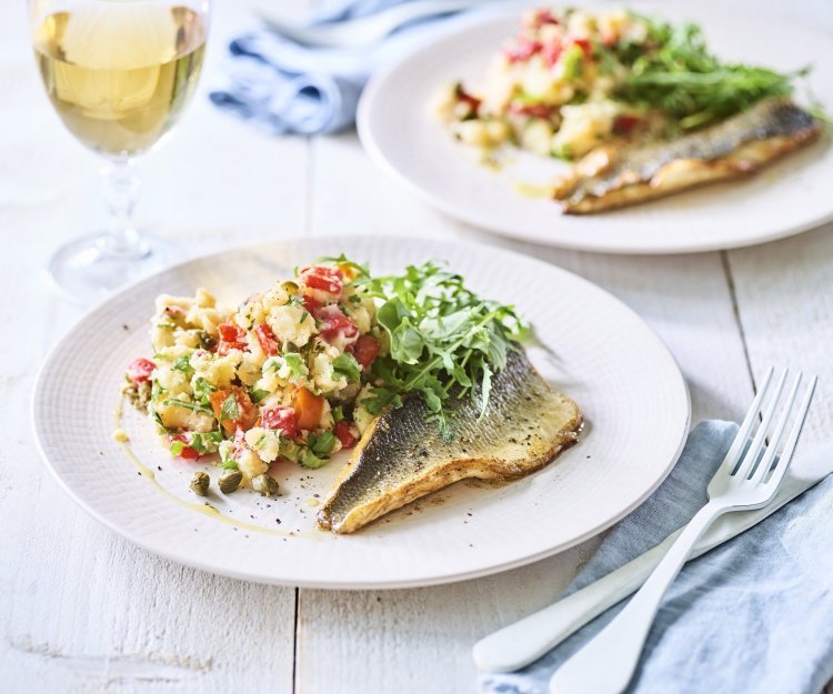 Zeebaarsfilet met een Zuiderse puree met geroosterde paprika, kappertjes en rucola Op tafel staan twee borden met het gerechtje op. Het is een aardappelpuree met stukjes paprika, kappertjes en rucolasla. En een krokant gebakken zeebaarsfilet. Er staat ook een glaasje witte wijn langs.