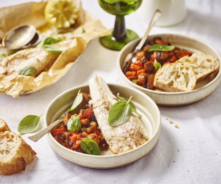 Zeeduivel uit de oven met caponata Twee bordjes gevuld met heerlijke homemade tomatensaus met olijfjes en kappertjes. Hier bovenop ligt het stukje vis en enkele blaadjes basilicum. Er staat ook een glaasje wijn op tafel met groene voet.