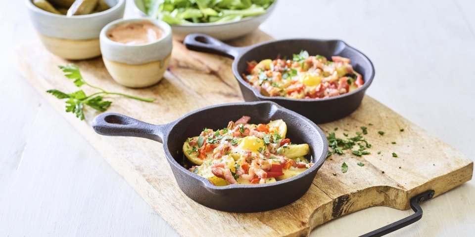 Aardappelplakjes met spek, paprika en kaas voor de gourmet Twee gourmetpannetjes staan te sieren op een houten snijplank. Als bijgerechtjes hebben we een groene salade, opgelegde augurkjes en een cocktailsausje.