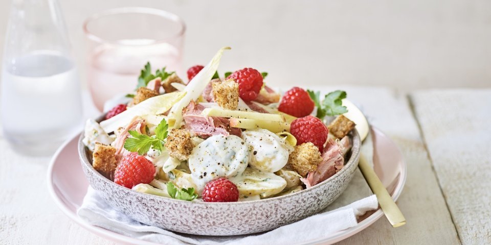 Aardappelsalade met Breydelham, appel en framboos Een rijkelijke aardappelsalade is geserveerd in een grote kom. Je ziet de aardappel met de dressing, stukjes Breydelham, framboosjes en appel. De kom staat op een pastelkleurig roos bord. Er staat ook een karaf water langs met een glas in dezelfde tint als het roze bord.