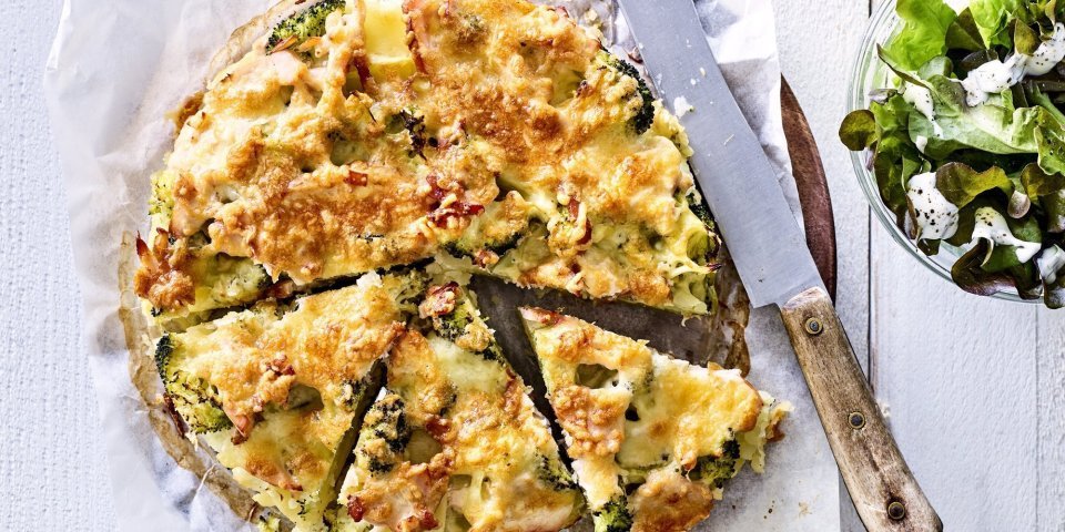 Aardappeltaart met broccoli Deze hartige taart lijkt op een quiche maar is toch anders. Deze is gemaakt van aardappelschijfjes, broccoli en kippenwit. Ze ligt op een houten ronde snijplank met boterpapiertje onder, ze is al voor de helft aangesneden. Rechtsboven staat nog een kommetje gemengde sla.