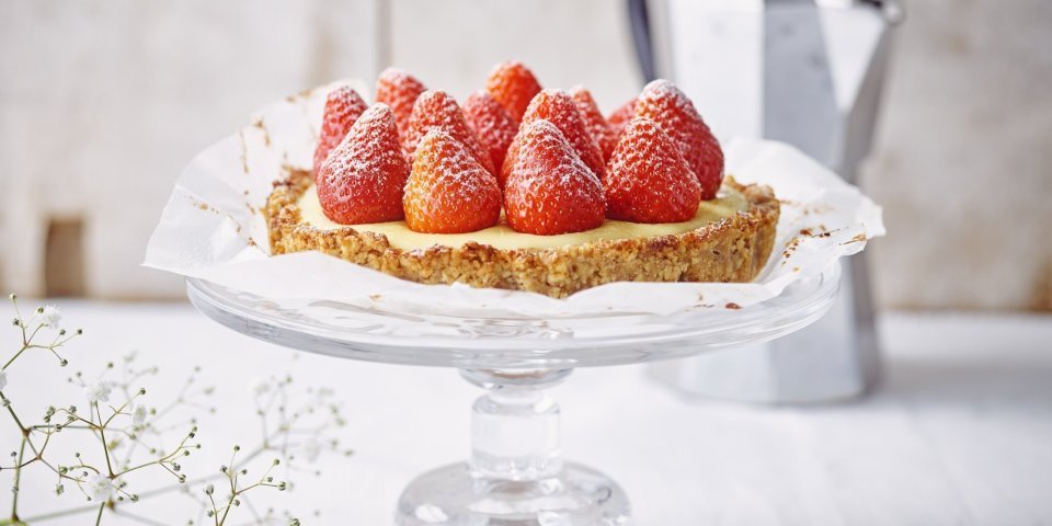 Aardbeientaart met vanillepudding en notenbodem De taart staat de stralen op een kristallen taartplateau. De verse aardbeien op de taart stelen de show. Achteraan de foto staat een kleine percolator om verse koffie te maken.
