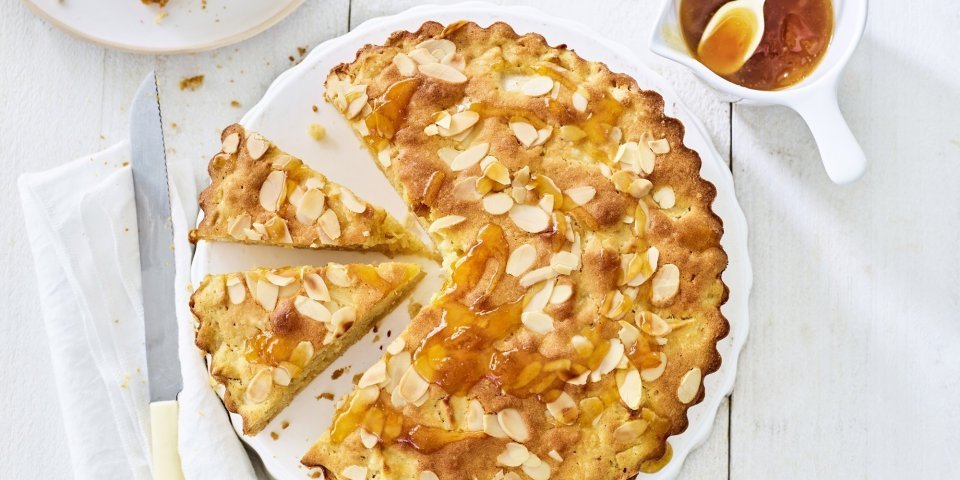 Goudgele appelcake staat op een houten witte tafel. De ronde cake is al voor een kwart aangesneden. Er is al een stukje uitgenomen en er staat een potje met abrikozengelei bij. 