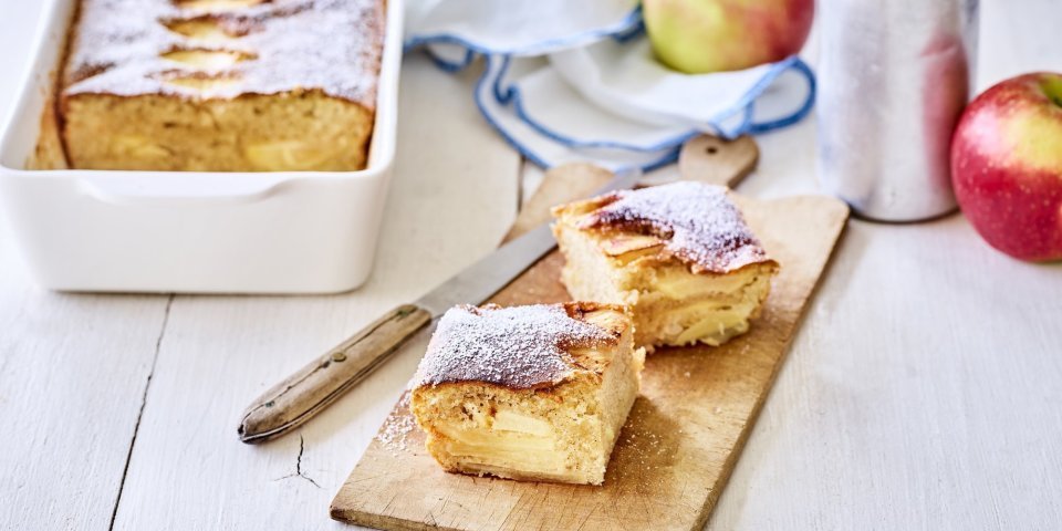 Een verse appelcake uit de oven, je reukt hem waarschijnlijk al. Er is een stukje uitgesneden en deze ligt opnieuw aangesneden op een houten plankje. Op tafel ligt een witte servette met blauwe randjes, nog enkele verse appels en de strooipot met bloemsuiker. 