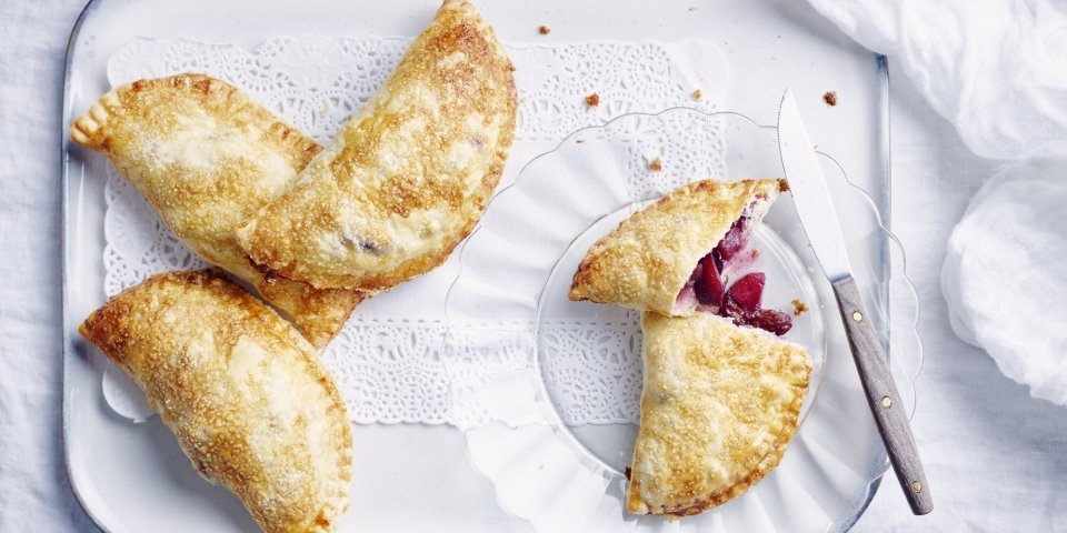 Appelflappen met blauwe bessen Er liggen 4 appelflappen klaargemaakt op een wit plateautje. Eén appelflap is opengesneden, door de combinatie te maken met blauwe bessen heeft de vulling mooi met appels een mooie rode kleur.