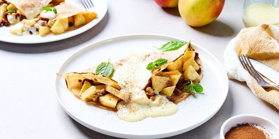 Appelstrudel-pannenkoek Op tafel staan twee borden met elk hierop een dichtgevouwen pannenkoek, die gevuld is met stukjes appel en rozijnen. Wij serveren hem met vanillesaus en verse blaadjes munt. Er liggen ook nog enkele appelen in de achtergrond en een restje vanillesaus.