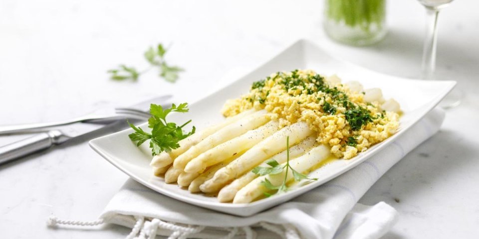 Op een rechthoekig wit bord liggen gekookte asperges, afgewerkt met geplette gekookte eieren, peterselie en gesmolten boter. Klaar om aan te vallen. 