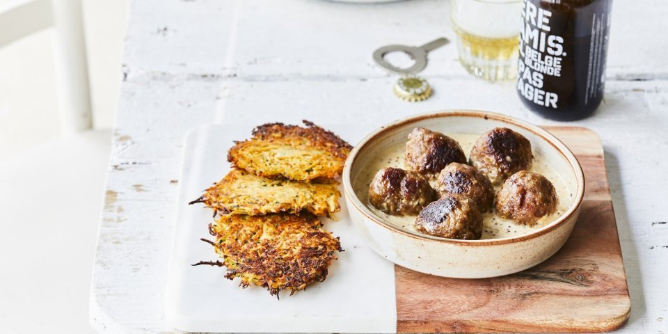 Gebakken balletjes liggen in een diep bord op de mosterdsaus, de krokante groenterösti's liggen er langs. Er staat een flesje bier op tafel met een klein glaasje.