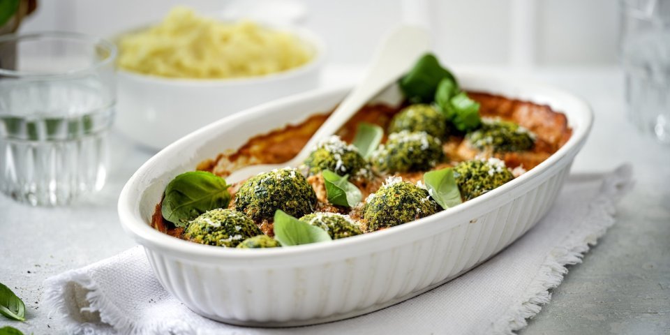 Balletjes van boerenkool in paprikasaus Een witte ovenschotel staat op tafel op een dikkere keukenhanddoek. De ovenschotel is gevuld met rode paprikasaus en hierop liggen de groene vegetarische balletjes van boerenkool. In de verte zie je ook nog een kommetje puree, klaar om aan tafel te gaan dus!