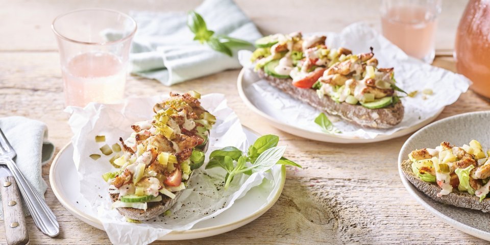 Belegd broodje met kruidige kip Er staan 3 bordjes klaar op een houten tafel. De ideale lunch om te benuttigen buiten in de zon. De halve broodjes zijn belegd met kruidige kip en verse groentjes.
