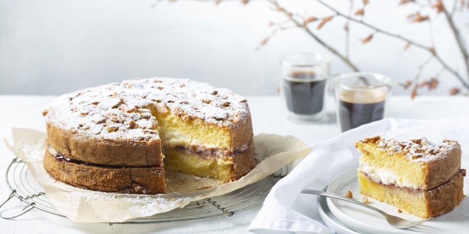 Een biscuit met vanillepudding en slagroom staat op tafel op een taartrooster. Er is al een stukje aangesneden om de lijnen duidelijk te laten zien van de pudding en slagroom. Koffie uitgeschonken, vorkje prikken en aanvallen die taart. 