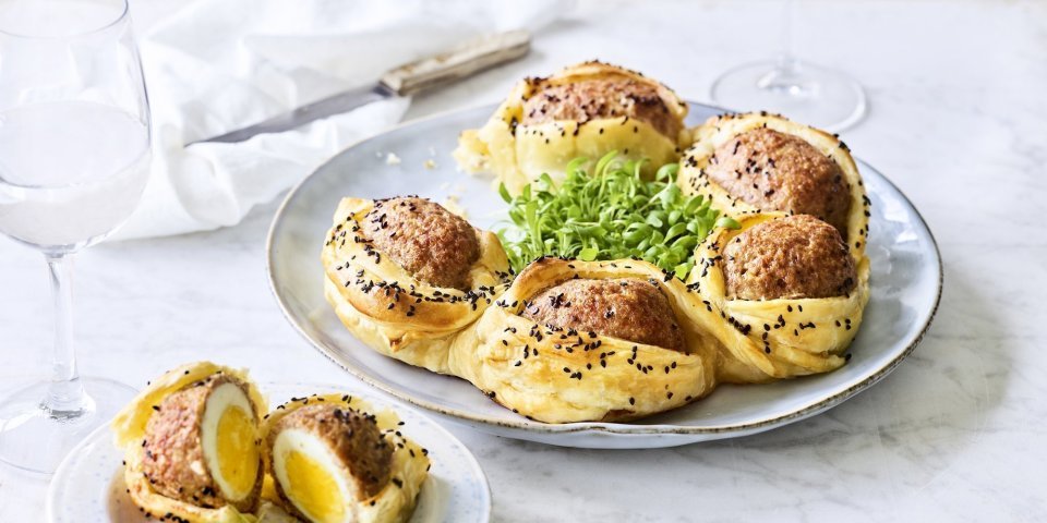 Bladerdeegkrans van vogelnestjes Op tafel staat een bord waarop een bladerdeegkrans ligt. Er zitten grote gehaktballen tussen gevluchten met hierin een gekookt eitje. Het bladerdeeg is afgewerkt met zwarte sesamzaadjes wat zorgt voor een leuke spikkeling.