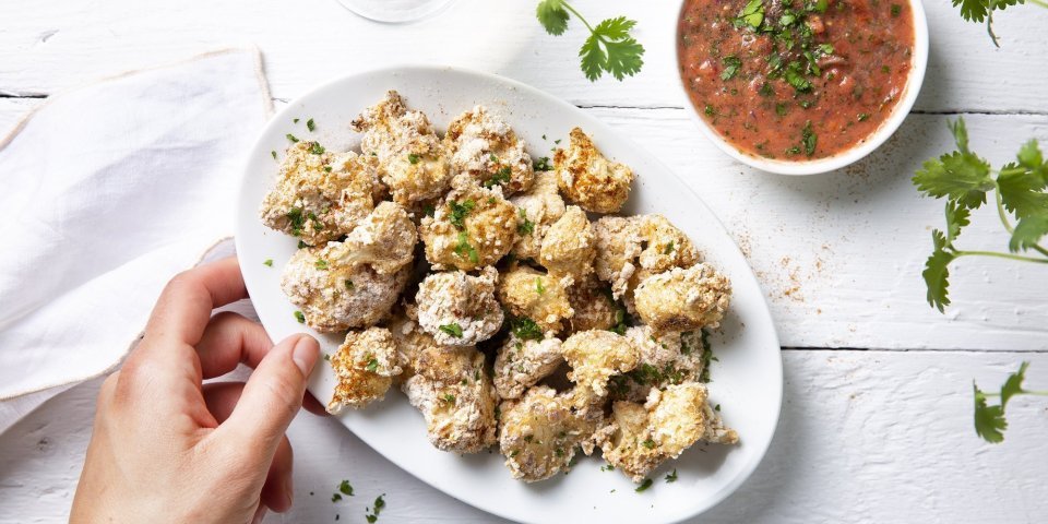 Bloemkoolbite met pittig sausje Krokante bloemkoolroosjes als aperitiefhapje, liggen klaar op een ovaal wit bordje. Er staat een pittige tomatensaus langs om te dippen. Tijd voor de apero te starten.