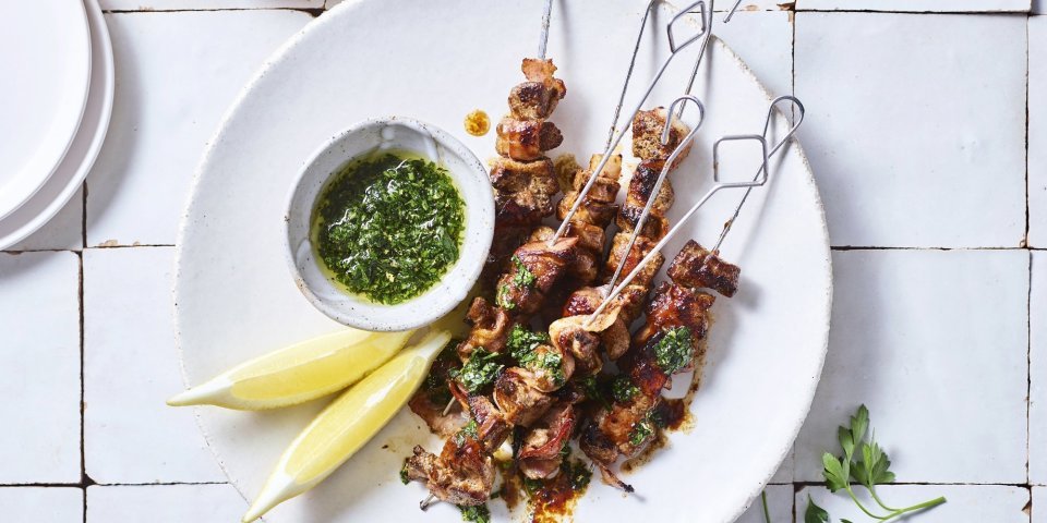 Brochette van lever en spek met kruidenolie Verschillende spiesjes liggen op een bordje te blinken, ze zijn geserveerd met een groene frisse kruidenolie en partjes citroen.