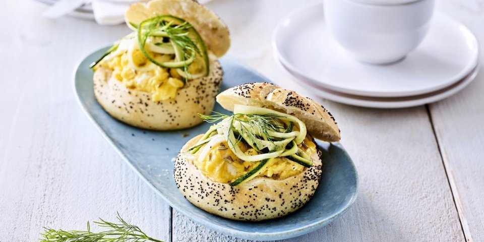 Twee pistolets liggen op een grijsblauw bordje. De pistolets zijn opengesneden en gevuld met roerei en courgetteslingers. 