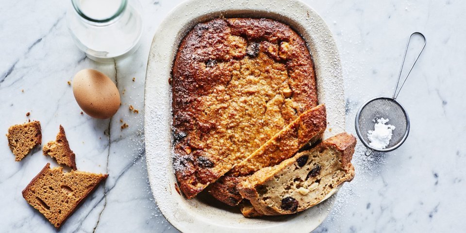 Een heerlijke homemade broodpudding staat op een marmer aanrecht in een witte ovenschaal. Hij is al aangesneden en een stukje uit de vorm genomen. Om de foto meer boddy te geven ligt er een eitje, flesje melk, stukje peperkoek en bloemsuiker naast.