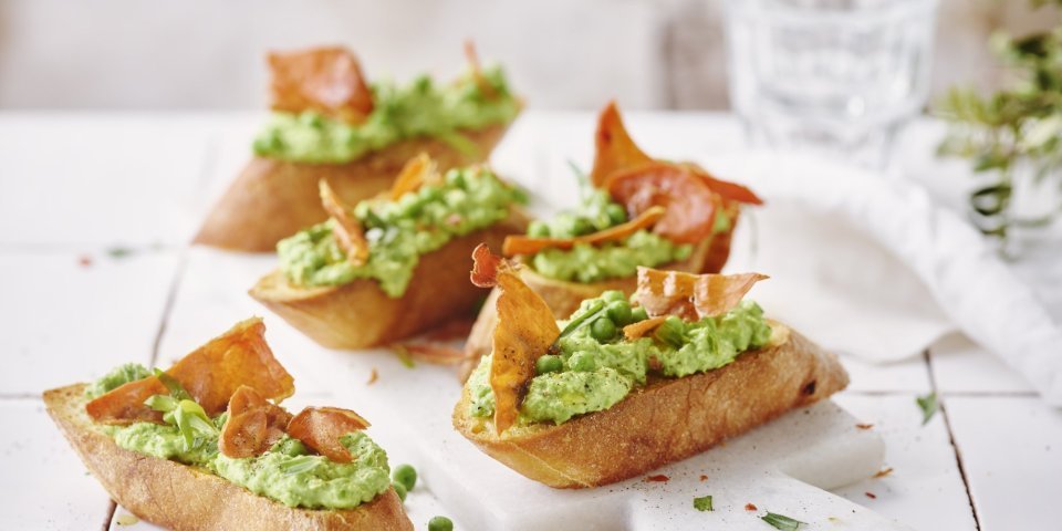 Bruschetta met geitenkaas, erwten, dragon en gedroogde ham Vijf klaargemaakte bruschetta liggen op tafel. De spread van geitenkaas en erwten kleurt mooi groen, de bruchetta werd afgewerkt met een stukje krokant gebakken gedroogde ham.