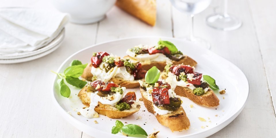 Bruschetta met paprika, burrata en pesto van basilicum met walnoten Op een houten, witte tafel staat een groot wit rond bord met hierop de bruschetta geserveerd. Ze zijn afgewerkt met extra blaadjes basilicum. Er staan ook kleine bordjes naast en de vijzel waarin de pesto is in gemaakt.