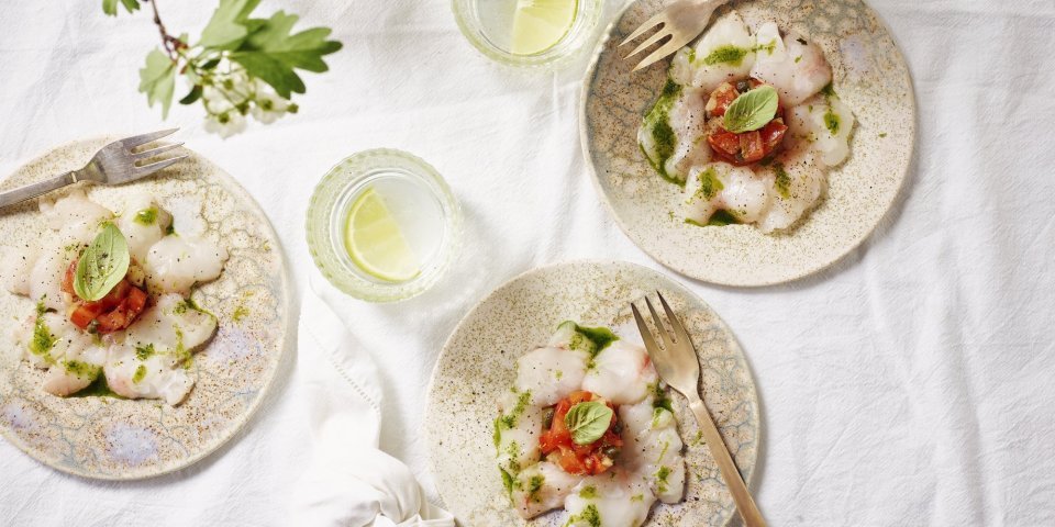 Carpaccio van zeeduivel met tomatentartaar en basilicumolie Drie feestelijke bordjes staan te schitteren op de tafel met een wit linnen tafelkleed. De vis is gesneden in fijne schijfjes en de tartaar van tomaten ligt in het midden van het bordje. Afgewerk met groene basilicum, heerlijke aperitiefhapje.