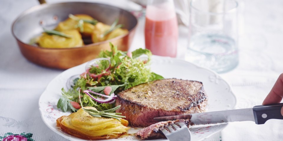 Chateaubriand met millefeuille van aardappelen en een kruidenslaatje met frambozendressing Een hoofdgerecht om U tegen te zeggen. We serveren een malse chateaubriand met millefeuille van aardappelschijfjes en knapperige kruidensalade. De frambozendressing staat langs het bord in een klein glazen flesje. Er staat ook een glas water boven het bord, naast peper en zout.