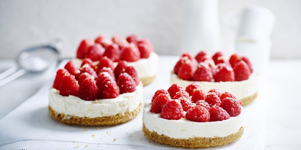 No bake-cheesecake met witte chocolade en frambozen Wij maakten 4 kleine cheesecakes en staan op een wit boterpapiertje. Ze zijn afgewerkt met verse frambozen die een mooi contast geven met het witte van de kaas.