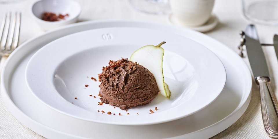 Chocomousse met een crumble van spek In een diep wit bord ligt een bolletje chocomousse met hierop de crumble van spek en een plakje peer. Bovenaan zie je een heerlijke kopje kofie staan. Perfect dessert om het feestmenu in glans af te sluiten.