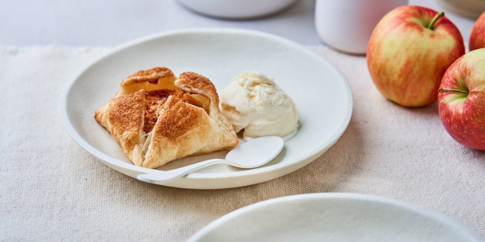 Op een mooi wit bord ligt een appeltaartje van bladerdeeg en een bolletje vanille-ijs. Er liggen ook enkele jonagold appelen langs het gerecht. 