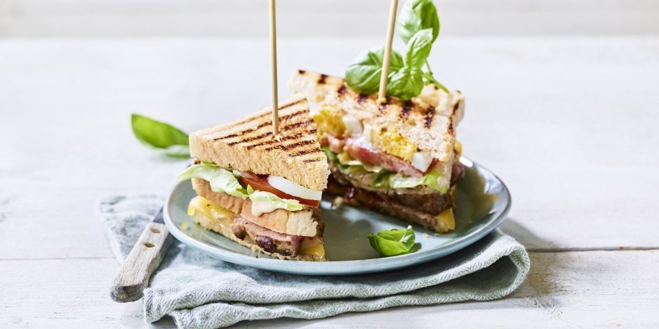 Club sandwich met gegrilde lamsfilet Goedgevulde club sandwich met lamsvlees en rauwkost. Gepresenteerd op een bord met middenin een prikker, klaar om op te eten.