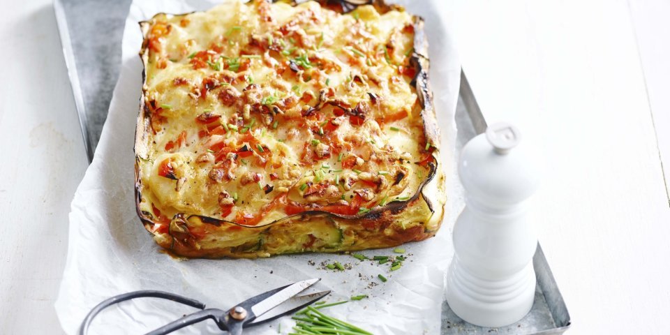 In een ovenschotel ligt een geweven taartje van courgette en aardappel met stukjes rode paprika. Hij heeft mooie kleuren van geel, rood en groen door de verse bieslook. 