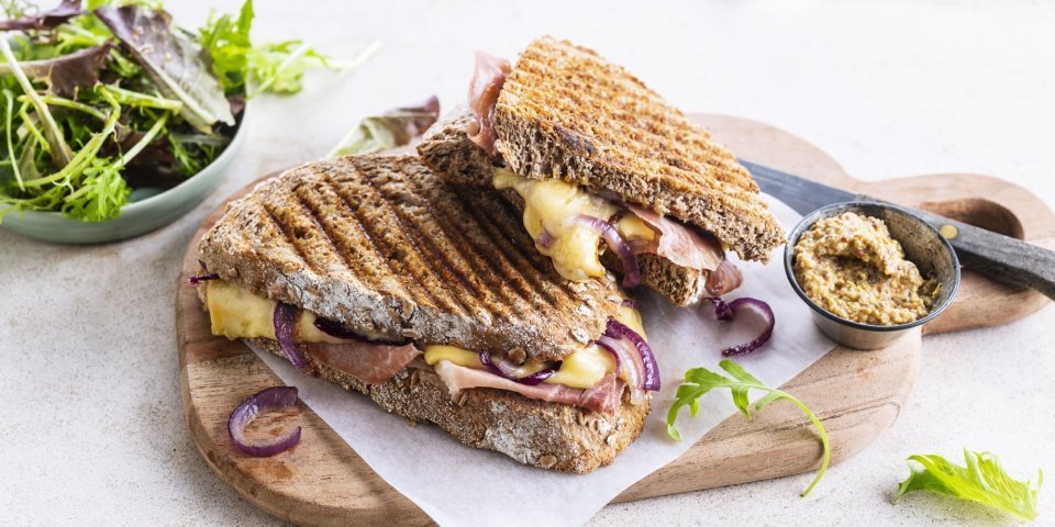 Croque met ham, rode ui en Passendale kaas De croque ligt op een houten plankje aangesneden. De kaas en rode ui zien er heerlijk uit. In de achtergrond zie je ook nog wat gemengde sla en een potje graantjesmosterd.