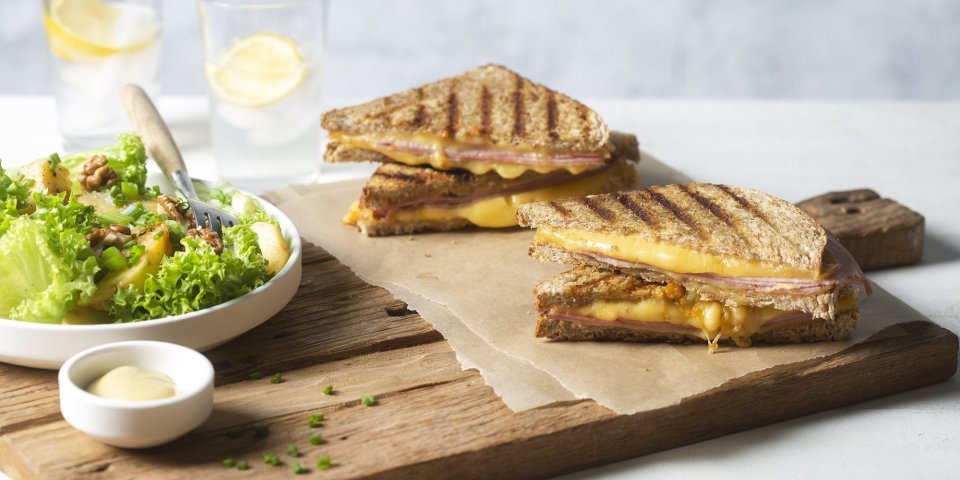 Croque monsieur met een salade van krulsla, appel en noten Een krokant gebakken croque monsieur ligt aangesneden op een houten snijplank. Lang staat een salade in een diep wit bord met een potje mayonaise voor te dippen.