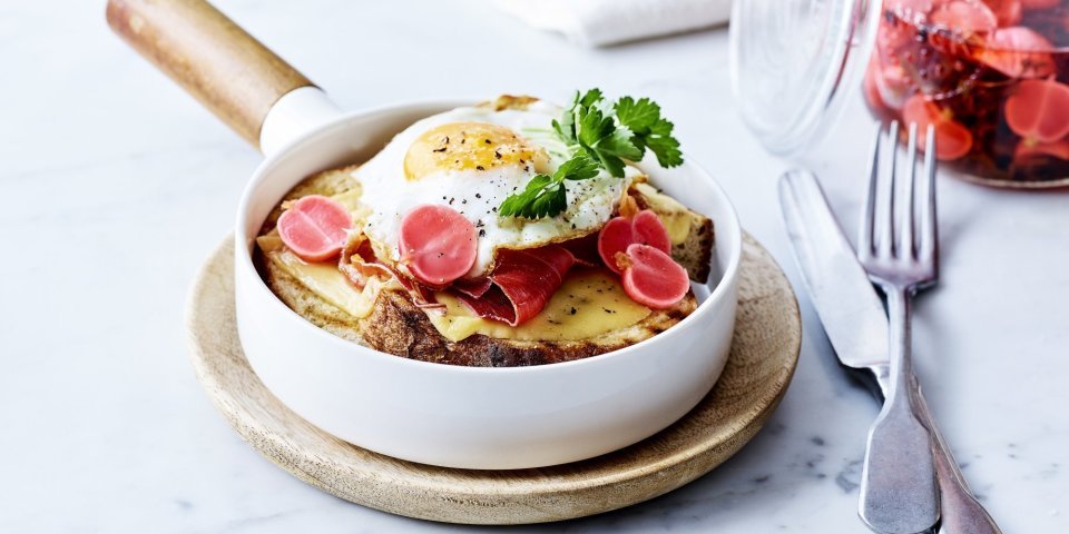 Croute au fromage met Averbode kaas, spiegelei en gepekelde radijs Croute au fromage met Averbode kaas in een witte pan, spiegeleitje er bovenop gepresenteerd met een takje verse peterselie. Op de achtergrond staan gepekelde radijzen in glazen pot.