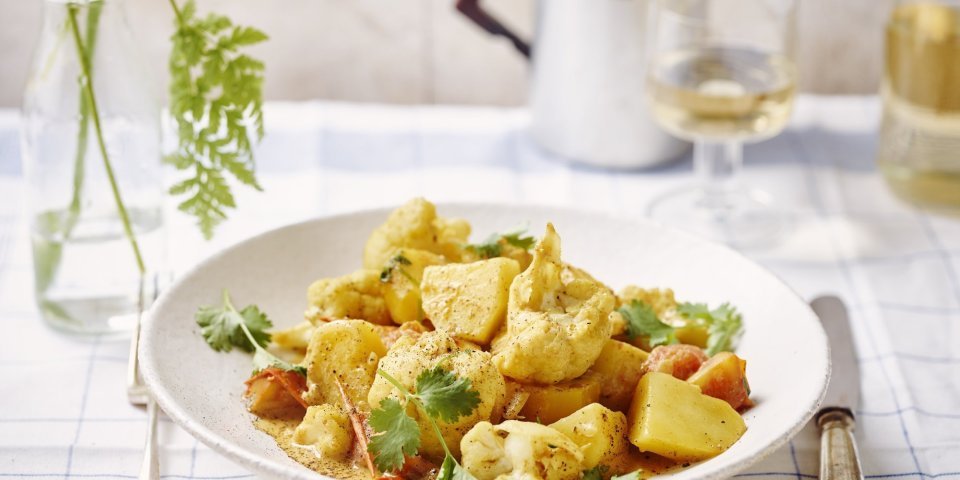 Heerlijke goudkleurige curry geserveerd in een diep wit bord met bloemkool en aardappelen. Er ligt bestek langsheen het bord en glaasje witte wijn. Ook zien we een vaasje met roze bloem en varentakje. 