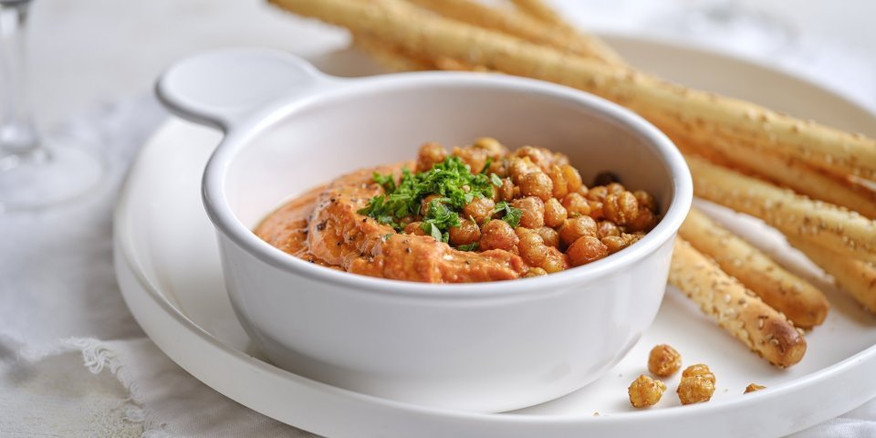 Dip van zoete puntpaprika met geroosterde kikkererwten Een wit porseleine potje is gevuld met een oranjekleurige tapenade. Het is er eentje van zoete puntpaprika met hierop geroosterde kikkererwten. Er liggen broodstengels langs om te dippen.