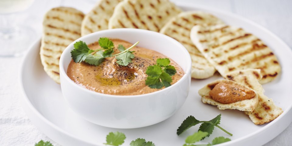 Dip van zoete puntpaprika met naanbrood Homemade paprikadip zit in een wit schaaltjes. Het naanbrood ligt er langs op een wit bord. Het gercht is afgewerkt met korianderblaadjes.