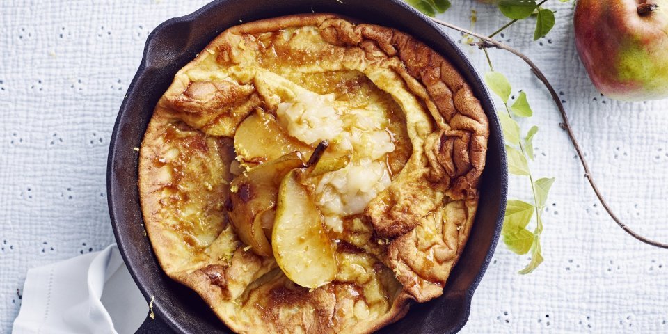 Dutch baby met gekarameliseerde peer en perencompote Een ovenpannenkoek met heerlijke peertjes, hij komt net uit de oven en ligt nog in de pan. Er liggen 2 verse peperen langs.