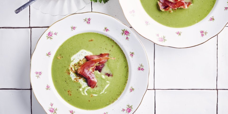 2 porties erwtensoep staan klaar op het werkblad van de keuken. De erwtensoep heeft een mooie groene kleur die mooi contrast geeft in de witte borden. De soep is afgewerkt met peterselieyoghurt, krokante ham en gefruite uitjes.