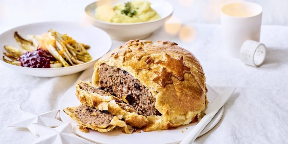 Rond gehaktbrood omhuld met bladerdeeg waarop sterren in bladerdeeg zijn teogevoegd. In het beeld liggen ook het gebraiseerd witloof met veenbessencompote en de puree. Kerstlampjes zien je flou in de foto.