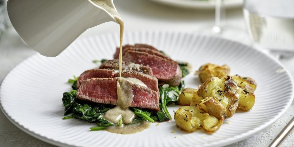Filet pur met blauwe kaas en gecrushte aardappelen Een wit bord staat op tafel met hierop de heerlijke krokante aardappel en de saignant gebakken filet pur op een bedje van spinazie. Er komt een sauskannetje in beeld die saus over het vlees giet. De tafel is gedekt voor twee met de perfecte maaltijd.