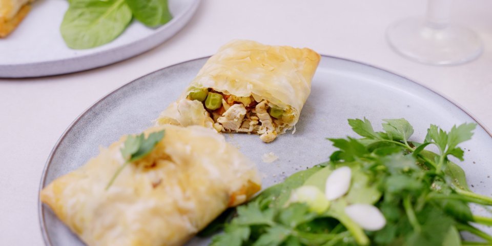 Filodeegpakje met kip en berloumi Een heerlijk cadeautje van filodeeg gevuld met stukjes kip, zoete aardappel, erwtjes en grilkaas ligt op een licht blauw bordje. We serveren het met een groen slaatje en lente-uitjes. Het filodeegpakketje is doormidden gesneden.