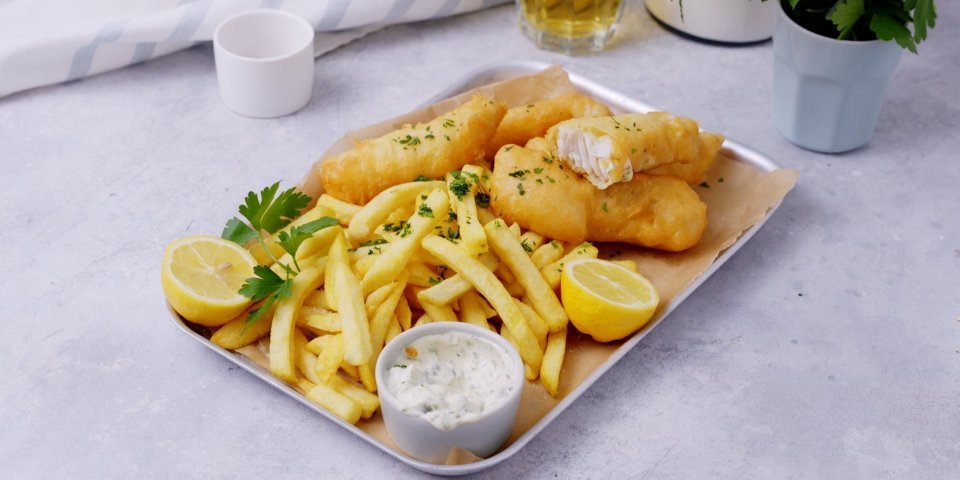 Fish and Chips Een metalen schotel is gevuld met homemade knapperige frietjes, fishsticks en verse tartaarsaus. Het gerechtje is afgewerkt met verse gesnipperde peterselie.