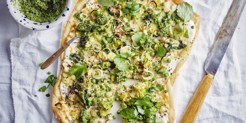 Op een witte snijplank ligt de rechthoekige flammkuchen. Hij heeft prachtige groene tinten vavn de erwtjes, prei en blaadjes waterkers. Er ligt een groot mes langs om aan te snijden een potje met extra pesto van waterkers.