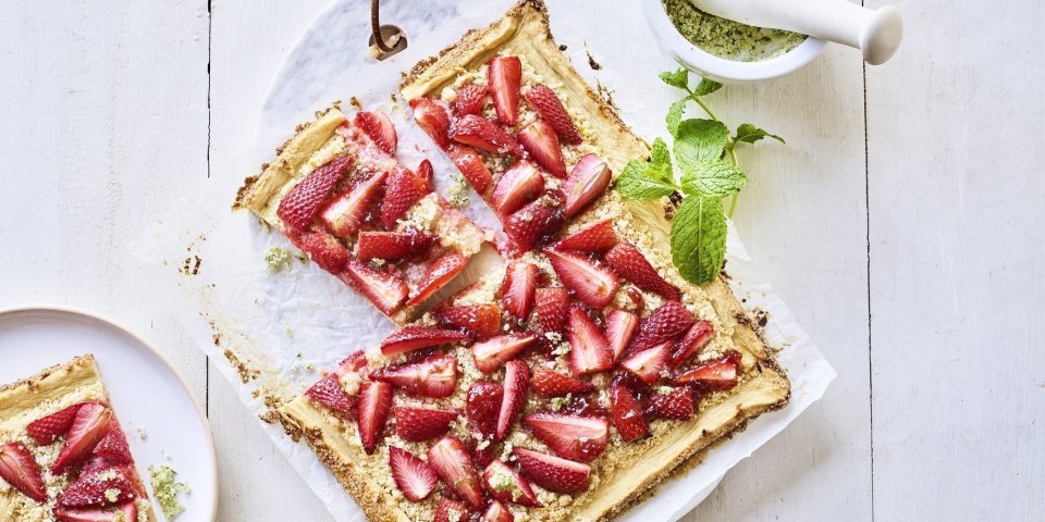 Een heerlijke plaattaart stat op tafel. Deze frangipanetaart ligt op een marmere snijplank en is afgewerkt met stukjes verse aardbeien en homemade muntsuiker. De taart is aangesneden en er ligt een stukje op een bord. 