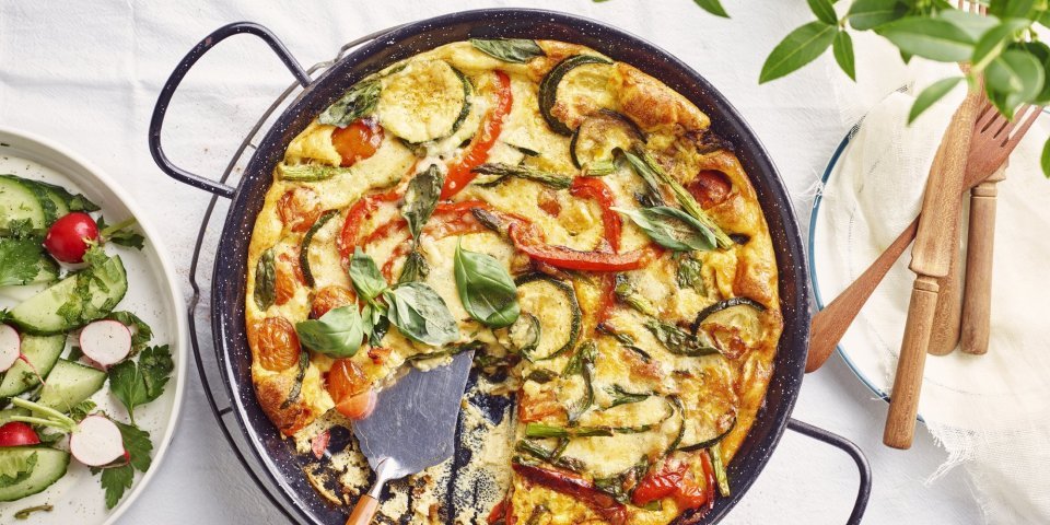 Frittata met kerstomaten, paprika, courgette en groene asperges De frittata staat op tafel in zwarte pan, waardoor hij nog mooier oogt dan dat hij al is. De tomaatjes, paprika, courgette en asperges geven leuke kleurtjes aan de frittata. Er staat een slaatje langs de linkse zijde, met komkommer, radijsjes en verse kruiden.