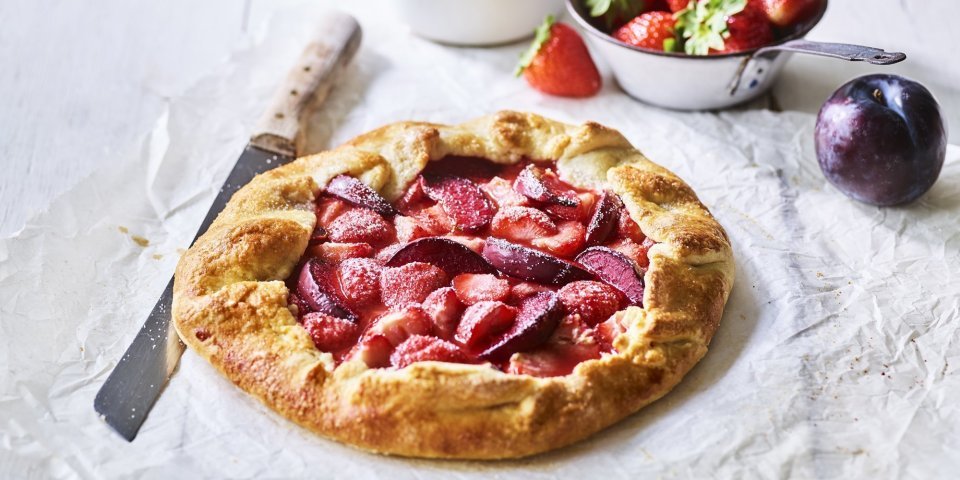 Galette met aardbeien en pruimen Een klaargemaakte galette staat op een houten witte tafel. Hij is prachtig goudgeel gebakken en het fruit oogt zoet-rode tinten. Er liggen nog wat aardbeien langs, alsook een paarse pruim.