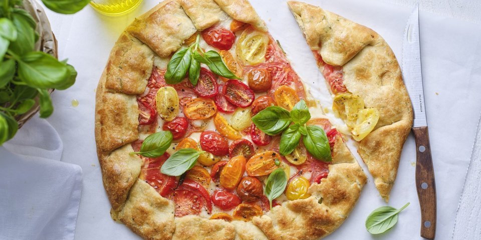 Galette met tomaten en mozzarella De galette van tomaten ligt op de tafel op een wit boterpapiertje. Hij is mooi gebakken en er is een stukje van gesneden. Er staat een basilicumplantje langs, alsook een flesje gouden olijfolie.