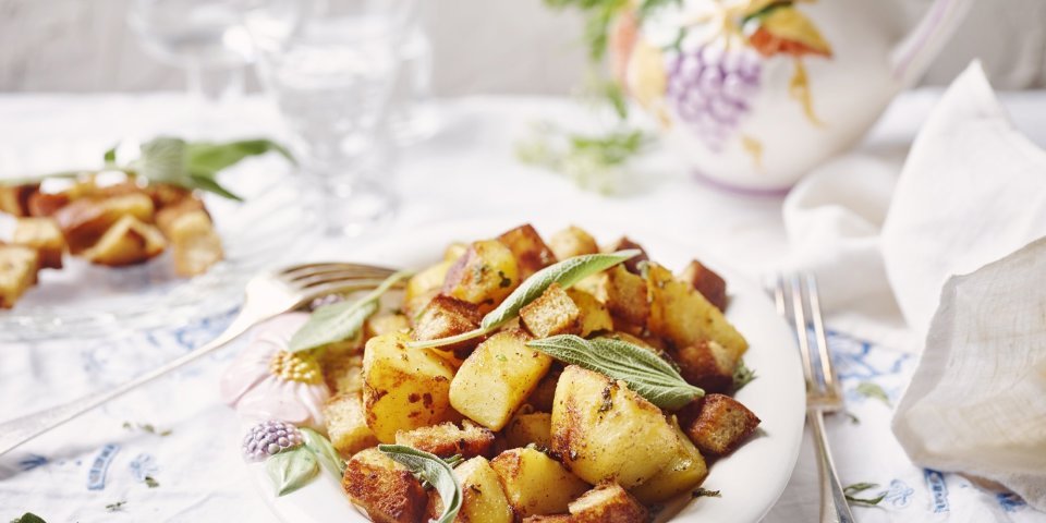 Gebakken aardappelen liggen in een groot wit bord op een gedekte tafel. De verse salieblaadjes geven de krokante aardappelstukjes extra kleur. 